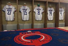 Locker room with five white striped soccer jerseys hanging in wooden cubby stalls, numbered 27, 15, 8, 14 and 4 above padded benches.