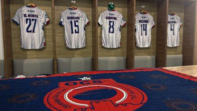 Locker room with five white striped soccer jerseys hanging in wooden cubby stalls, numbered 27, 15, 8, 14 and 4 above padded benches.