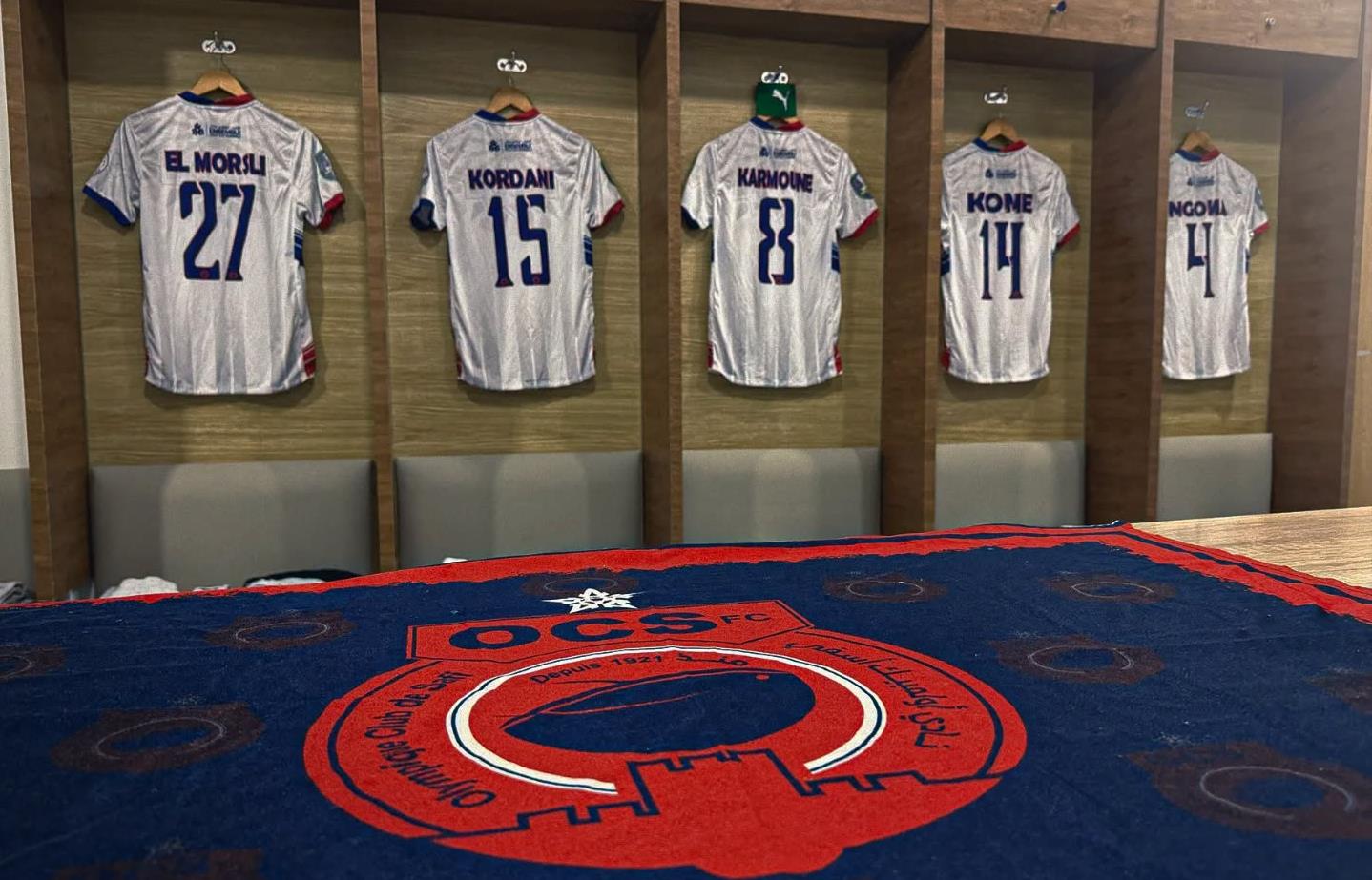 Locker room with five white striped soccer jerseys hanging in wooden cubby stalls, numbered 27, 15, 8, 14 and 4 above padded benches.