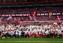 Soccer team in white shirts poses with trophy on the pitch as fans hold red banners in the stands.