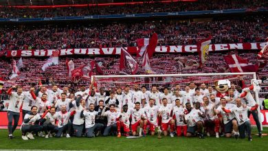 Soccer team in white shirts poses with trophy on the pitch as fans hold red banners in the stands.