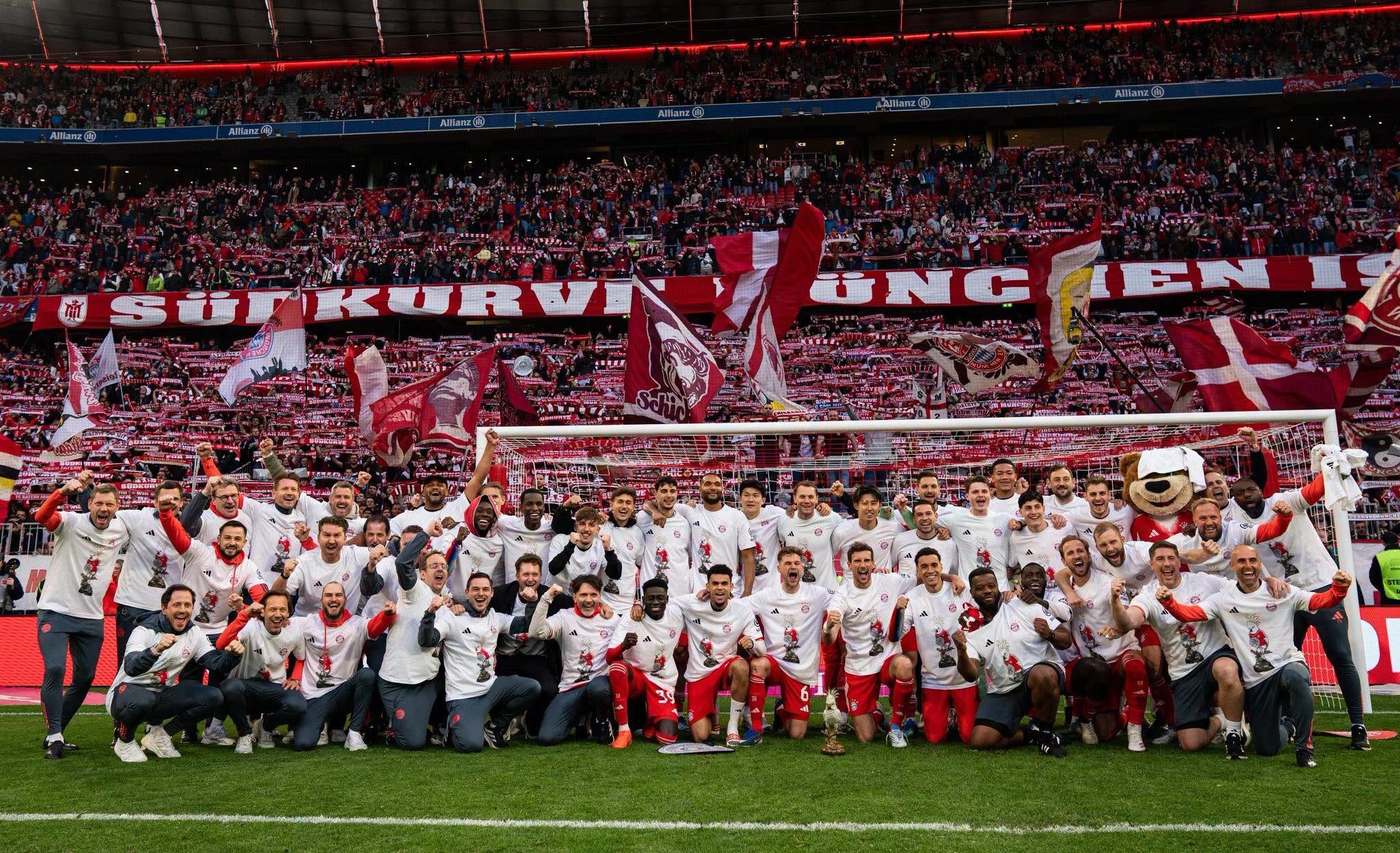 Soccer team in white shirts poses with trophy on the pitch as fans hold red banners in the stands.