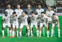 Eleven soccer players in white kits pose for a team photo on a green field with a stadium crowd behind them, goalkeeper in gray center back.
