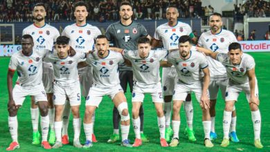 Eleven soccer players in white kits pose for a team photo on a green field with a stadium crowd behind them, goalkeeper in gray center back.