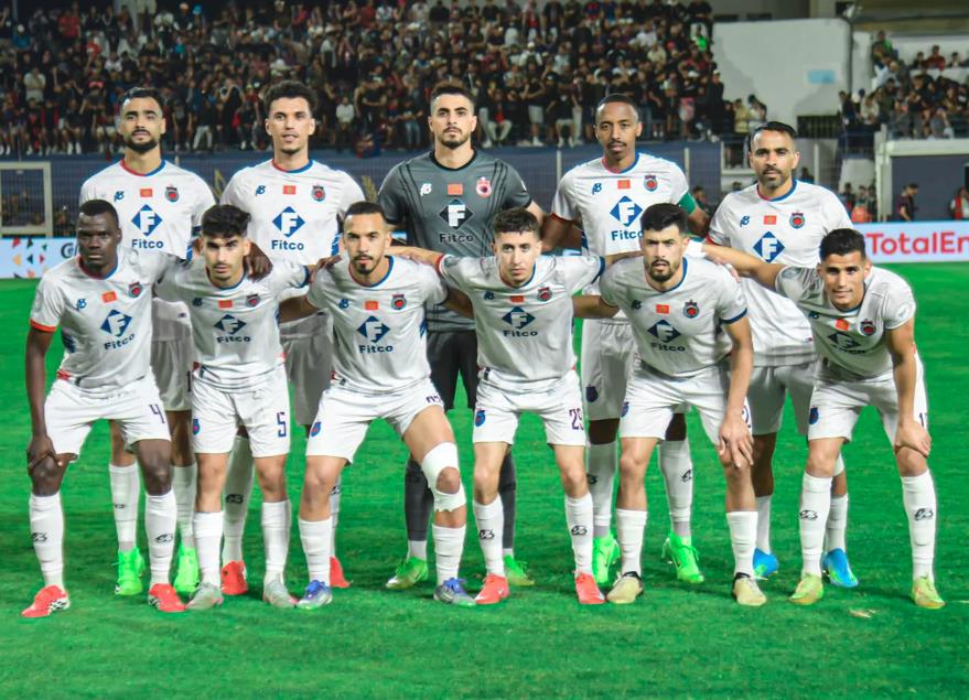 Eleven soccer players in white kits pose for a team photo on a green field with a stadium crowd behind them, goalkeeper in gray center back.
