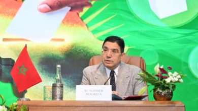 Panelist at a wooden desk with Moroccan flag, nameplate reading M. Nassir Bourirat, a water bottle, and flowers on the table with a bright green backdrop.
