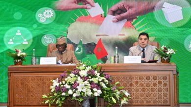 Two men sit at a carved wooden desk during a formal signing ceremony, flags of Pakistan and Morocco displayed between them, with a floral arrangement in front and a bright green backdrop showing hands exchanging a document.