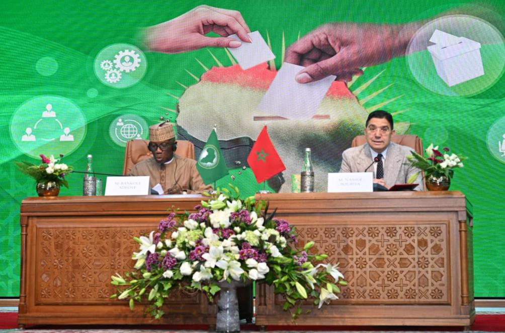 Two men sit at a carved wooden desk during a formal signing ceremony, flags of Pakistan and Morocco displayed between them, with a floral arrangement in front and a bright green backdrop showing hands exchanging a document.