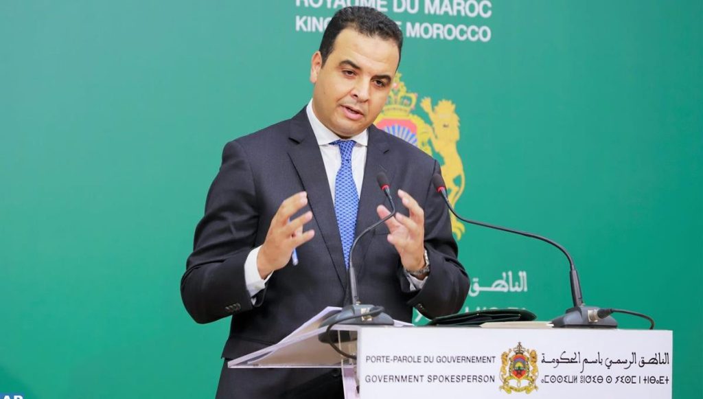 vis_0421202612374018 | MCG24 | موقع إلكتروني مغربي شامل متجدد على مدار الساعة Male official in a suit speaks at a podium during a government briefing, green backdrop with Moroccan emblem behind him.