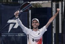 Male padel player in a white shirt and cap raises arms in victory, holding a paddle above his head.
