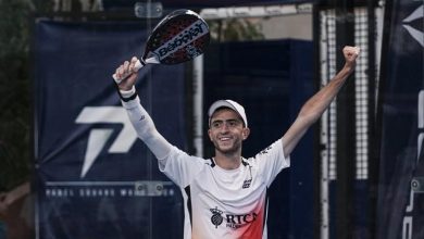 Male padel player in a white shirt and cap raises arms in victory, holding a paddle above his head.
