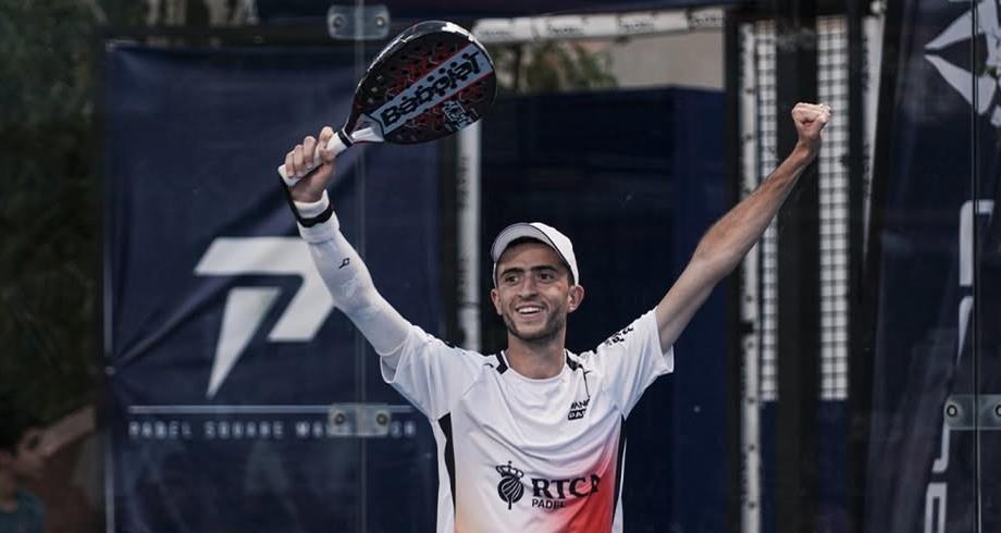 Male padel player in a white shirt and cap raises arms in victory, holding a paddle above his head.