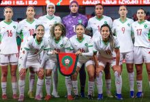 Morocco women's national football team posing for a pre-match group photo, holding a green-and-red shield banner on the field.