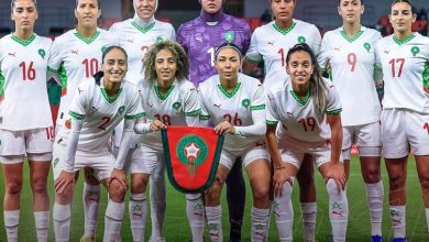 Morocco women's national football team posing for a pre-match group photo, holding a green-and-red shield banner on the field.