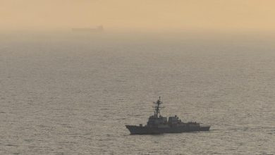 A gray naval destroyer sailing on calm water with a hazy orange sunset horizon behind it.