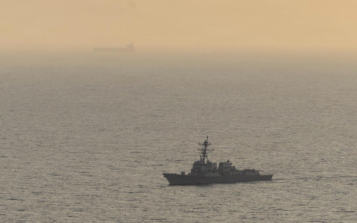 A gray naval destroyer sailing on calm water with a hazy orange sunset horizon behind it.