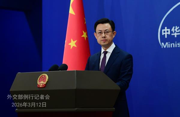 Man in a suit speaks at a podium with microphones during a press briefing, with a Chinese flag and blue Ministry backdrop behind him.