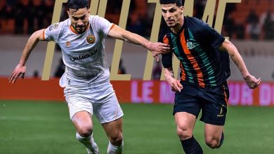 Two soccer players sprint on the pitch, one in white, the other in dark jersey with orange stripes, competing for the ball as a crowd watches.