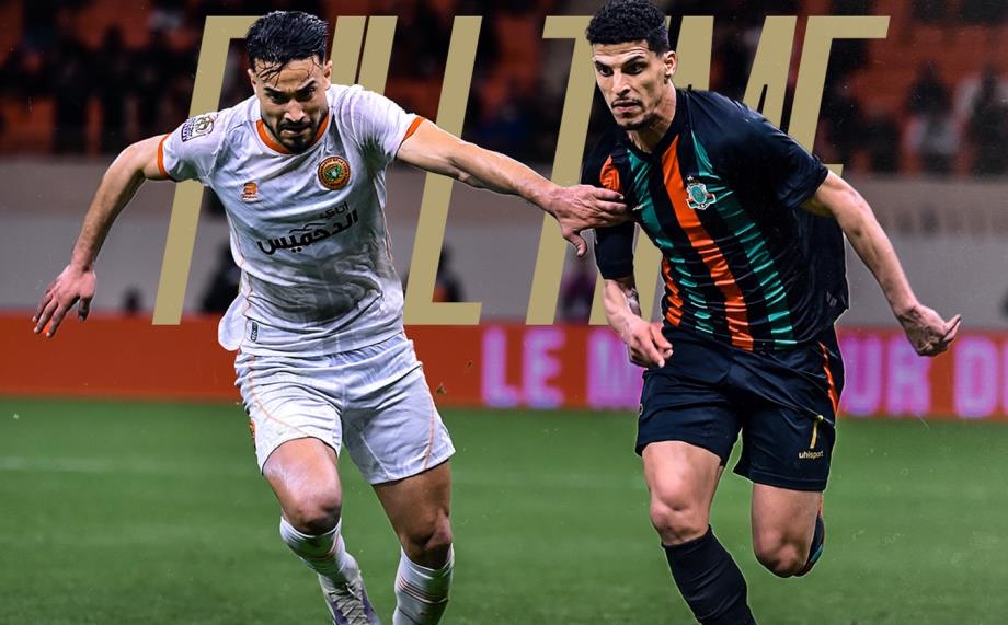 Two soccer players sprint on the pitch, one in white, the other in dark jersey with orange stripes, competing for the ball as a crowd watches.