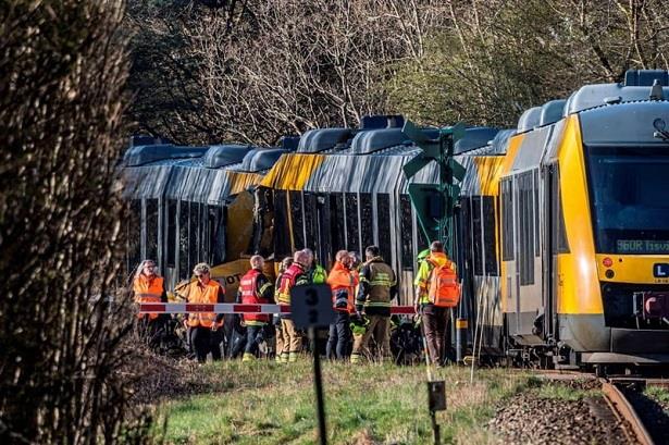 Emergency responders in high-visibility jackets inspect a derailed yellow passenger train on a track beside a wooded area.