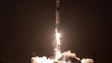 Nighttime rocket launch with a bright flame, exhaust plume, and rising smoke around a tall rocket.