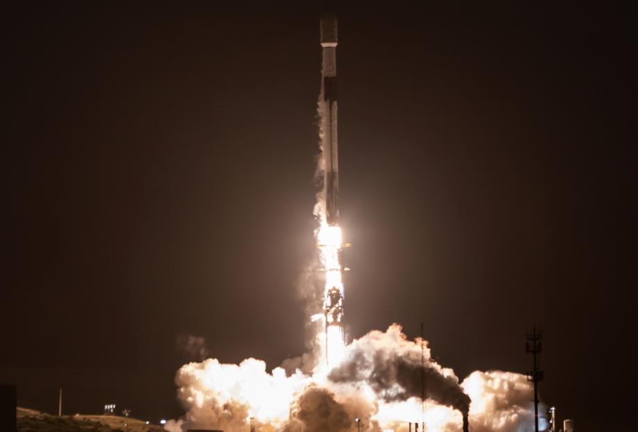 Nighttime rocket launch with a bright flame, exhaust plume, and rising smoke around a tall rocket.