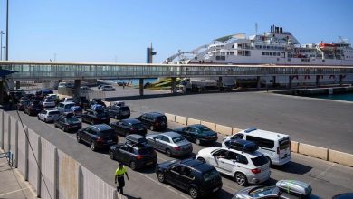 Port scene with a large white ferry docked, a queue of cars along the road, and a pedestrian in a high‑visibility vest near the terminal.