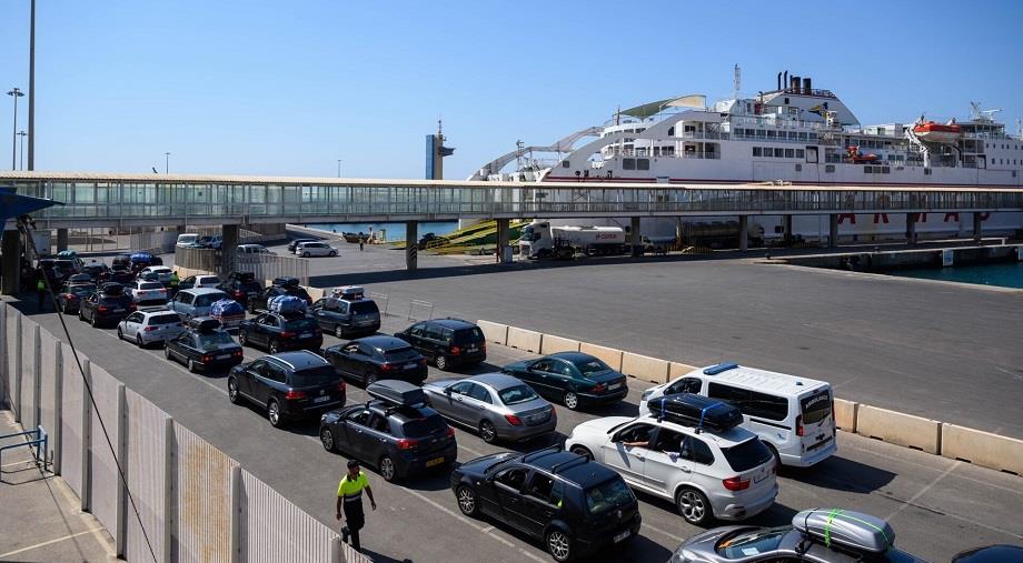Port scene with a large white ferry docked, a queue of cars along the road, and a pedestrian in a high‑visibility vest near the terminal.