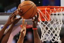 Hands of players contesting a basketball near the hoop during a game, with the net in the background and ball at the center of the action.