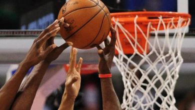Hands of players contesting a basketball near the hoop during a game, with the net in the background and ball at the center of the action.