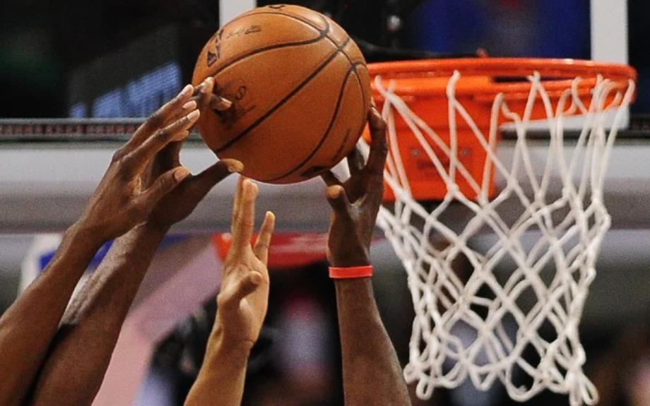 Hands of players contesting a basketball near the hoop during a game, with the net in the background and ball at the center of the action.