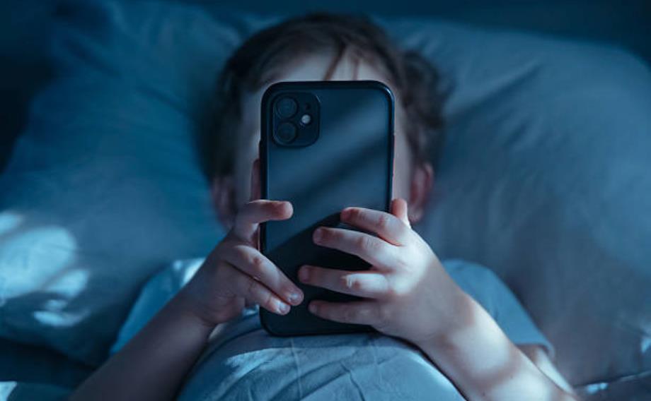 Child lying in bed at night, holding a smartphone up to their face with blue-tinted light