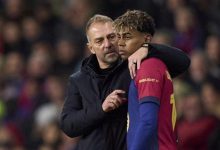 Coach with arm around a young footballer during a match, in a stadium crowd behind them.