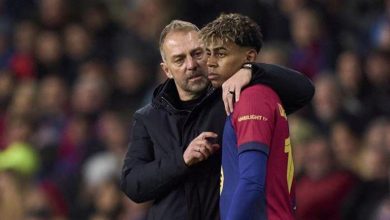 Coach with arm around a young footballer during a match, in a stadium crowd behind them.