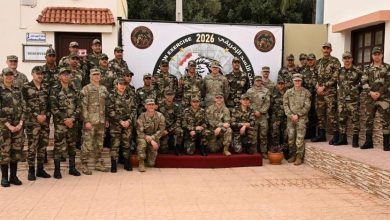 Group of soldiers in camouflage uniforms pose for a photo in front of a banner marked 2026 during a military exercise.