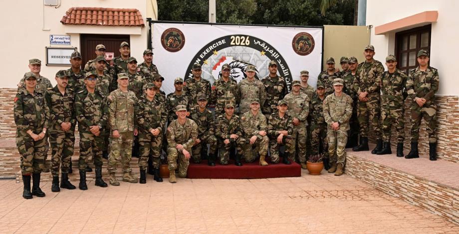 Group of soldiers in camouflage uniforms pose for a photo in front of a banner marked 2026 during a military exercise.