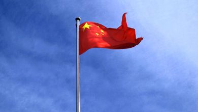 Red Chinese flag with yellow stars waving on a tall silver flagpole against a clear blue sky