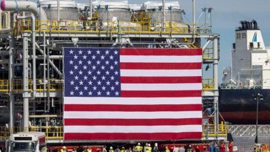 Large American flag hung over an industrial shipyard with workers and heavy piping in the background and a ship nearby.