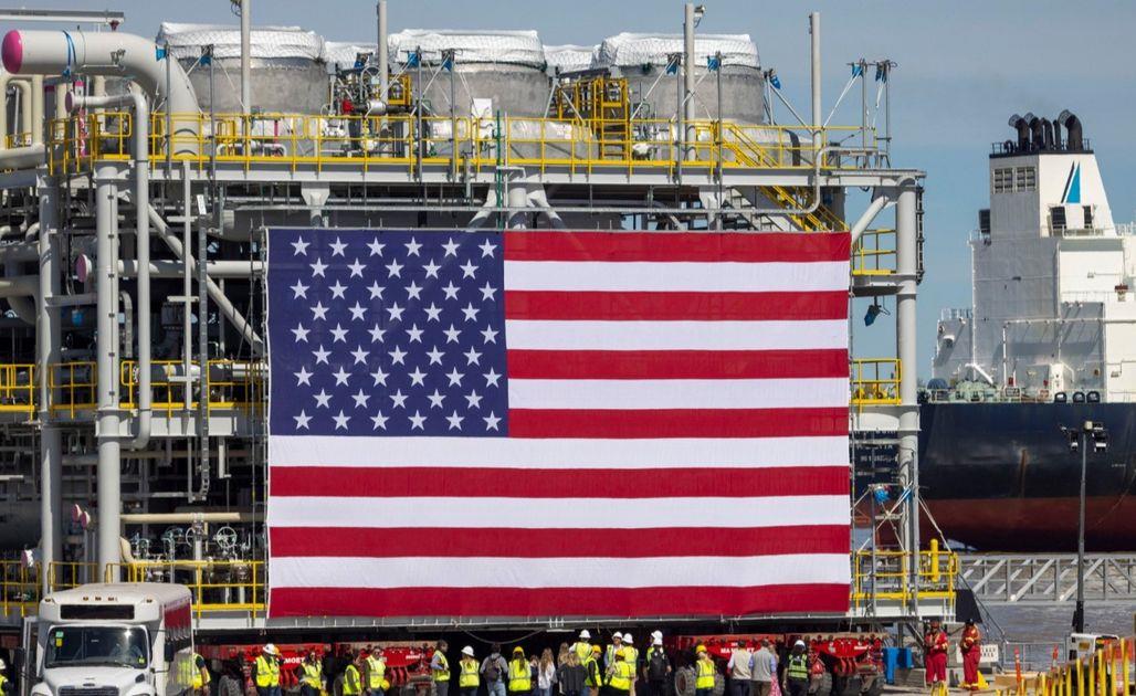 Large American flag hung over an industrial shipyard with workers and heavy piping in the background and a ship nearby.