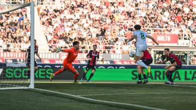 Soccer player in white number 8 shoots toward the goal as a red-clad goalkeeper dives to save, crowded stands in the background.