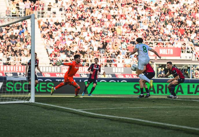 Soccer player in white number 8 shoots toward the goal as a red-clad goalkeeper dives to save, crowded stands in the background.