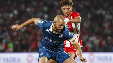 Soccer player in a blue kit dribbles the ball while a red-and-white defender closes in from behind on the field.