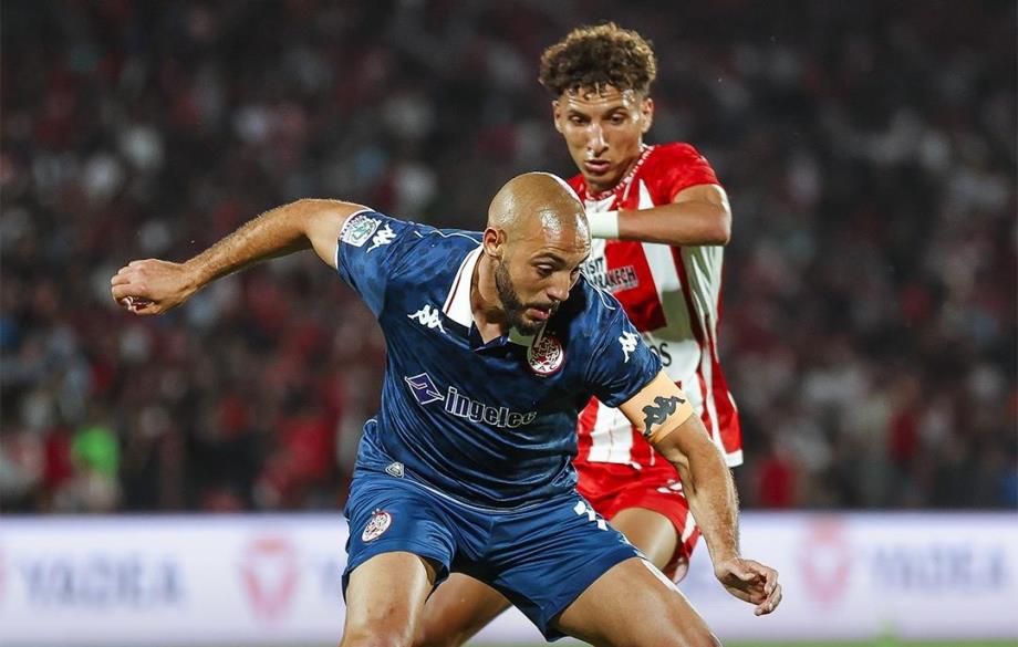 Soccer player in a blue kit dribbles the ball while a red-and-white defender closes in from behind on the field.