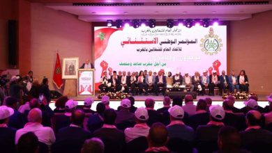 Formal conference on a stage with a panel of speakers, Moroccan flag, and a large screen with Arabic text behind them; audience seated in a hall.