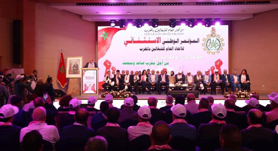 Formal conference on a stage with a panel of speakers, Moroccan flag, and a large screen with Arabic text behind them; audience seated in a hall.