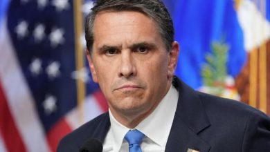 Man in a dark suit and blue tie speaking at a press conference, with the American flag in the background.