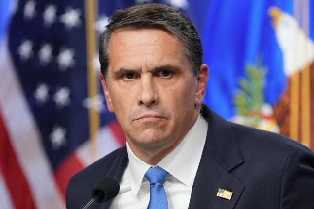 Man in a dark suit and blue tie speaking at a press conference, with the American flag in the background.