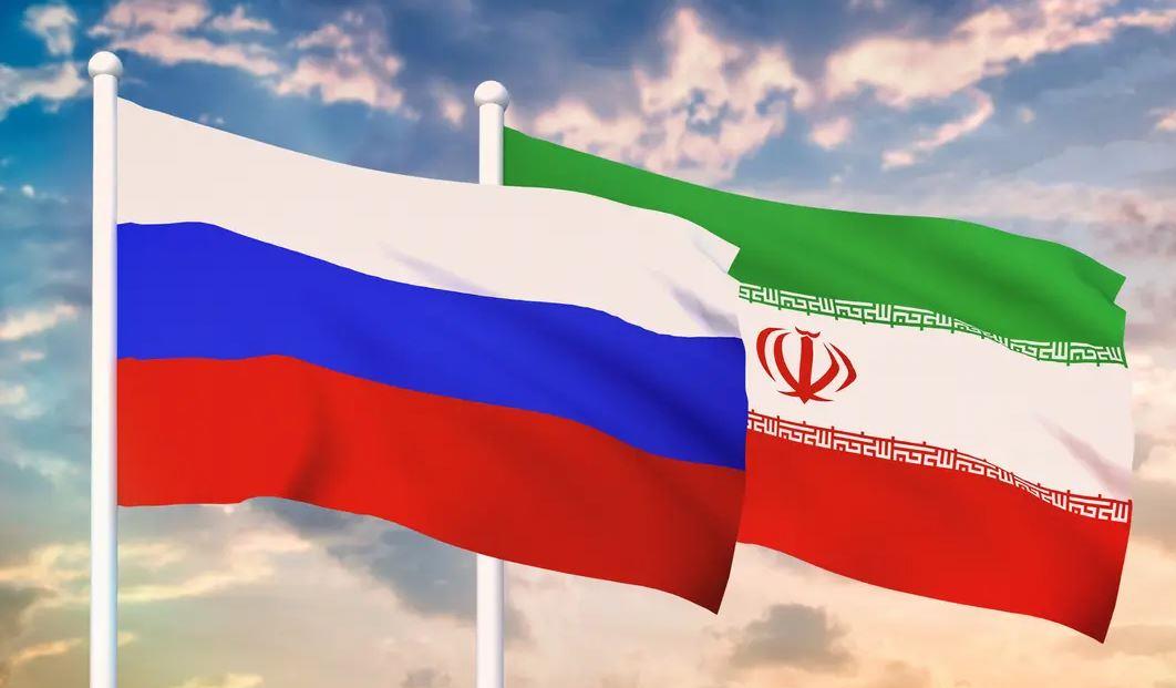 Russia and Iran national flags waving side by side against a cloudy sky on flagpoles.