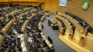 Wide view of a large conference hall with curved rows of desks filled with delegates, a speaker podium at the front, and a green emblem on the wall.