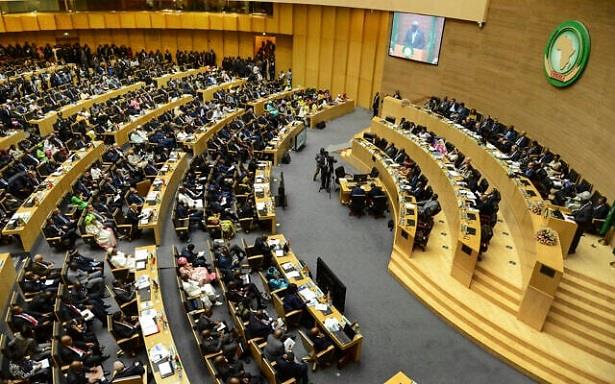 Wide view of a large conference hall with curved rows of desks filled with delegates, a speaker podium at the front, and a green emblem on the wall.